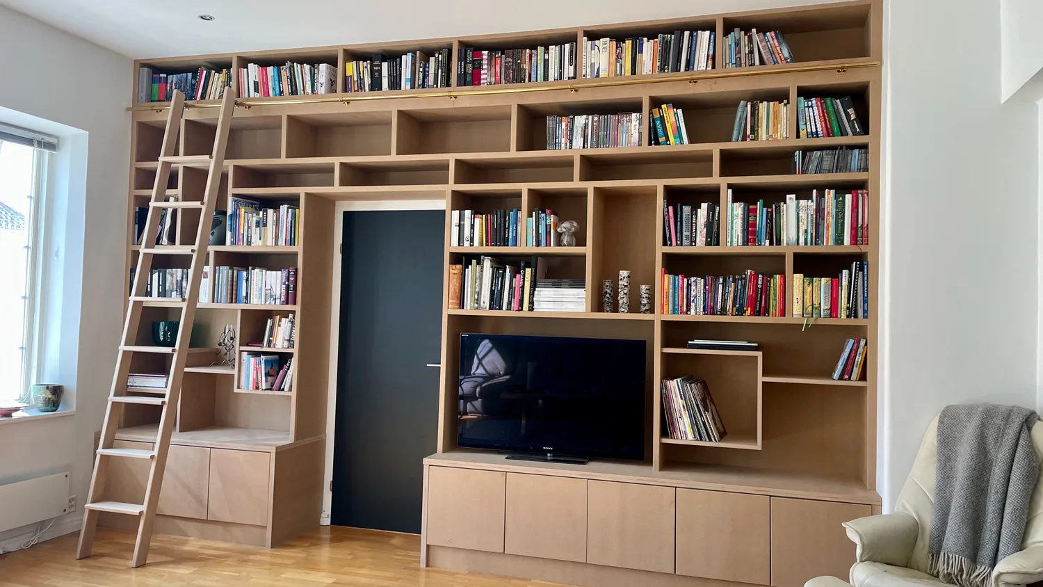 Vegghøy bokhylle som dekker hele veggen, med biblioteksstige til venstre, innfelt dør i midten og TV-benk.