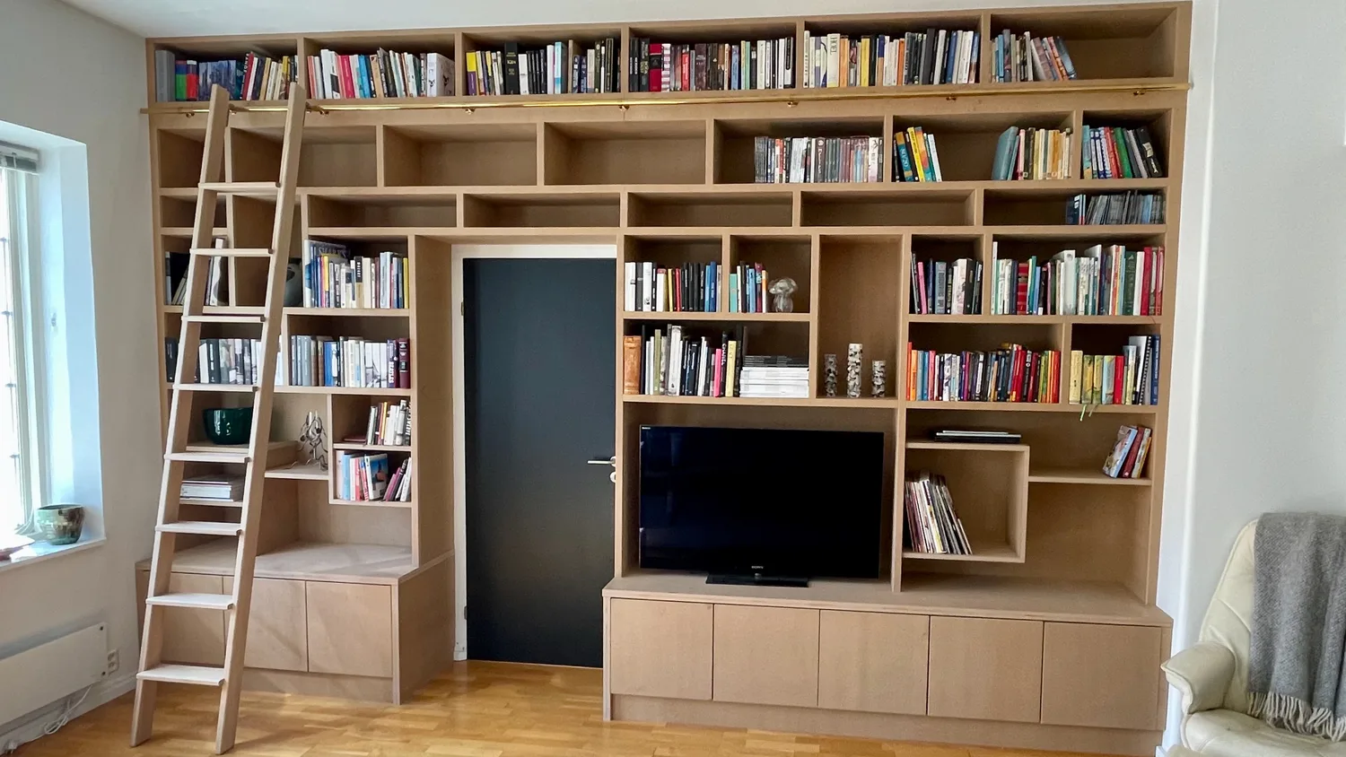 Hele bokhylla sett på skrå, med stige, innfelt dør og TV-benk.